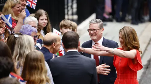 TOLGA AKMEN/EPA-EFE/REX/Shutterstock Prime Minister Keir Starmer and his wife Victoria Starmer in red dress with arms outstretched greet well-wishers as they arrive at Downing Street in London