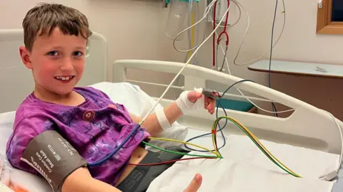 Stanley is lying in a hospital bed with cables connected to him and a machine. He is smiling at the camera.