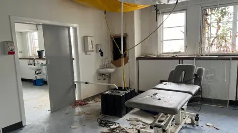 Image shows picture of a treatment room, with a sink in the corner and treatment bed in the centre of the room. 
Rubble has fallen onto the floor and bed, there is a hole in the wall close to the sink and a plastic tarpaulin covers the ceiling. 