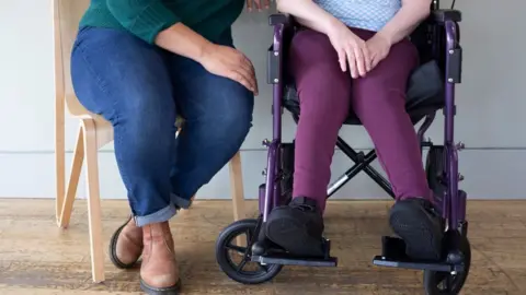 Two people sitting close together, One of the women has quadriplegic cerebral palsy and is a wheelchair user. The woman sat on the left is wearing a green jumper, blue jeans and brown boots, sat on a wooden chair, the person using the wheelchair has purple trousers and a blue top. They are pictured from the waist down.