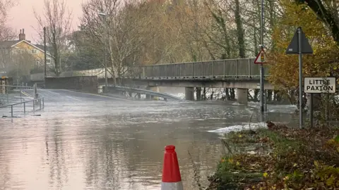 Harriet Heywood/BBC Flood water has taken over a road which can no longer be seen. Towards the back of the image the road reappears as it goes over a bridge. To the right of the image is a sign for Little Paxton. The sunrise is pink in the sky.