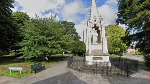 A Google Streetview of St Mary's Square. There is a large ornate monument in a park area, as well as a bench, lots of trees and a blue street sign. 