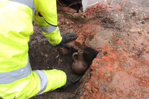 Mola A man wearing hi-viz protective clothing bends over a hole in the ground which contains a brown Bartmann jug