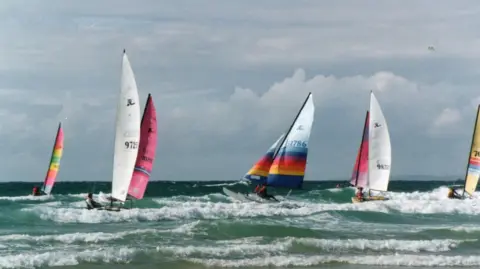 Royal Channel Islands Yacht Club A series of Hobie Cats sail in green surf and waves. The sky is cloudy behind them.