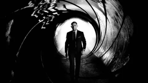 Daniel Craig as James Bond in the opening titles. He is wearing a black suit and walking towards the camera. 