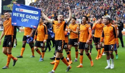 PA Media Wolves footballers, wearing old gold and black kits, on the pitch at Molineux on a lap of honour after winning promotion. Two of the players are holding a blue flag saying we're going up