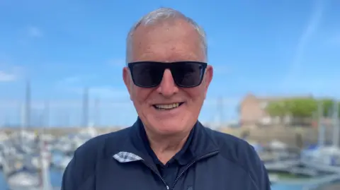 A man with grey hair in a blue jacket. There are boats in a marina behind him.