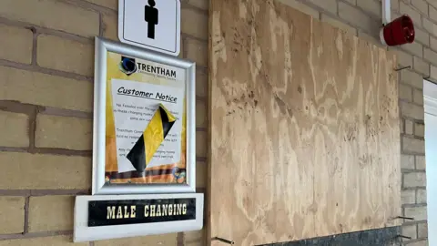 Ann Gannon / BBC A boarded?up doorway in a brick corridor with a large sheet of plywood covering the entrance. A sign above reads “Male Changing”, with a printed customer notice from Trentham Community Sports Centre taped nearby, partly obscured by black?and?yellow hazard tape. A male?toilet symbol hangs above the notice, and a red alarm device is fixed to the wall on the right.