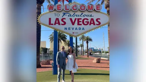 Cancer Reseach UK handout Sam and Liv Shaw standing by the Welcome to Fabulous Las Vegas Nevada sign in their wedding outfits. The road is lined with palm trees. They have their arms around each other and are smiling. It is a sunny day. 