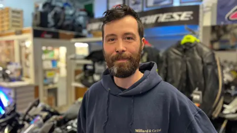 Clinton Millard - A man with black hair and a black beard. He has a blue hoodie on.