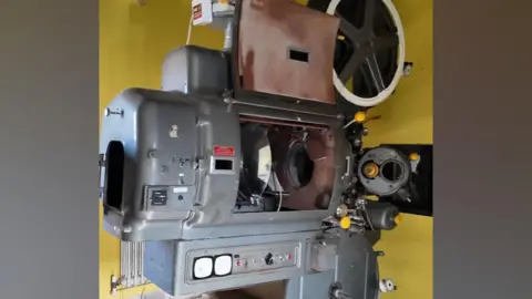 Nostalgia Nerd A large grey film projection machine is shown in a small room with yellow walls, with a central panel open to show some of its internal machinery.