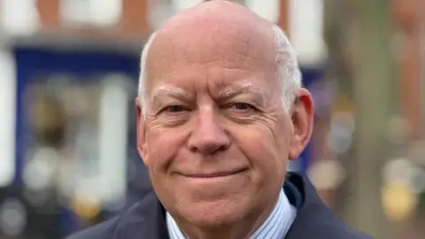 A man is smiling at the camera. He has white hair. He's wearing a blue and white shirt and blue jacket.