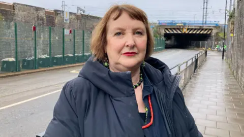 Angela Souter, a woman with short dark hair wearing green and black jewellery and a dark padded jacket, standing in front of a road under a bridge