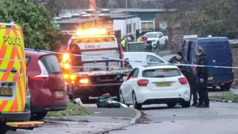Dawid Wojtowicz/ BBC Scene in a residential street with police vehicles, a breakdown truck, various cars, and houses in the background