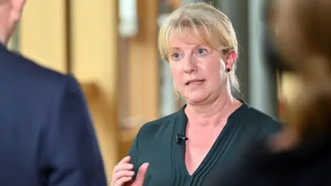 Shona Robison, with blonde hair and wearing a green dress, speaks to reporters in the Scottish Parliament 