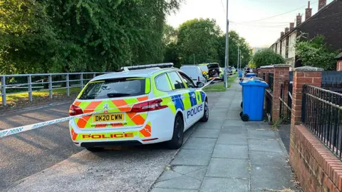 Merseyside Police vehicles inside a cordon with a row of houses on the right of Tobruck Road in Huyton and trees to the left lining the road on a sunny evening.