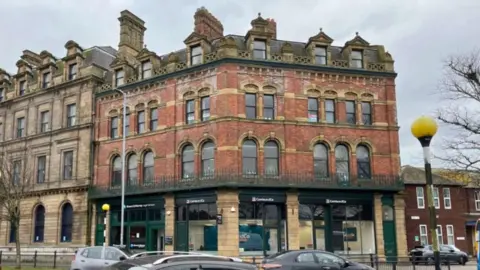 A four-storey brown brick building with shop fronts on the ground floor. Several cars are driving by.