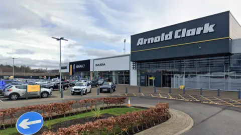 Arnold Clark headquarters in Glasgow. Cars sit in the forecourt and there is a large grey building in the background.