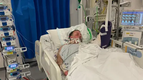PA Media A picture shows Jason Knight in hospital, hooked up to many different machines and monitors