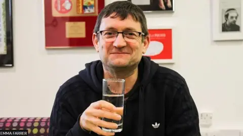 Emma Farrer Paul Heaton sitting holding a drink of clear liquid. He is wearing glasses and smiling. There are pictures on the wall behind him