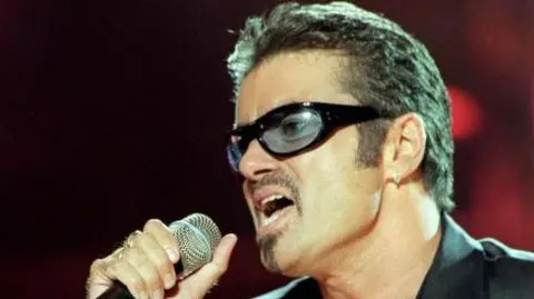 PA Media George Michael performing for NetAid at Wembley Stadium. He wears tinted glasses and has a short goatee beard.