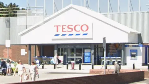 People walk outside a white fronted Tesco superstore which has black bollards in front of it.