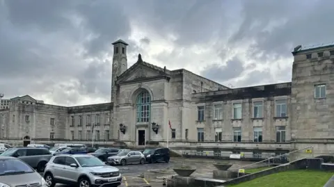 Southampton City Hall - a large white stone building with a central grand entrance with a large arched window above the door. In the foreground is a car park full of cars.