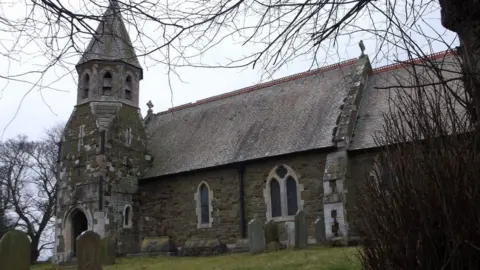 A grey and green sandstone church building with a short square tower topped by an octagonal tiled roof. Bare branches of trees overhang the churchyard with headstones.