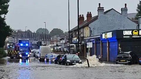  West Midlands Fire Service Water is seen gushing from the ground, with water completely flooding a road in the foreground while in the background is a fire engine parked beyond the flood. There is a street with two rows of house and a line of parked cars visible.