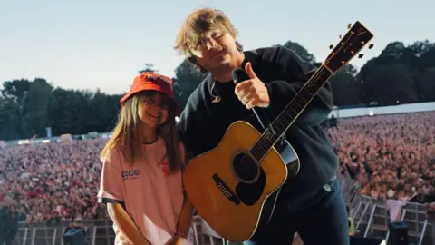 Evie-Mae stood with Lewis Capaldi on stage in front of a large crowd. He has his thumb up and has a guitar in front of him. Evie is wearing a red bucket hat. 