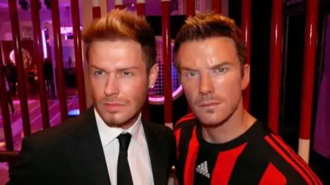 Andy Harmer A man, Andy Harmer, stood next to a waxwork figure of David Beckham, wearing a black and red AC Milan kit.
