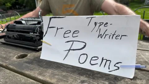 A close-up of Patrick's "Free typewriter poems" sign. It is handwritten on a piece of paper which is folded in half, and being held down by two pens attached on either side. There is also an old, black typewriter in the background on a wooden table.