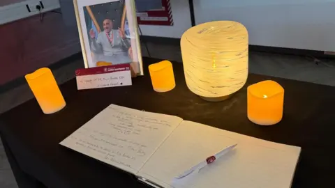 A book of condolence in honour of the late Sir Billy Boston is open with a white pen with the Wigan Warriors logo on it. The book is on a table with a black tablecloth and a framed photograph of the rugby league legend with candles.
