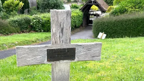 A wooden cross in the ground, with a plaque on it. Behind it is a hedge and a covered gateway into the graveyard.