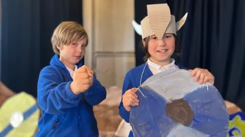 Sam Dixon-French/BBC Two children pretending to be vikings. The one on the left is blonde and has his fists raised and the one on the right is wearing a viking helmet made of cardboard and a painted homemade shield