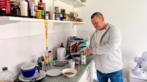 Martin Giles/BBC Gary James is wearing a grey hooded sweatshirt with a white T-shirt underneath. He is opening a carton of milk while standing in front of some kitchen units, about to make a bowl of cereal and a cup of tea