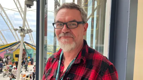 A man wearing glasses, with a beard and short grey hair. He is wearing a red plaid shirt and standing in front of a window.