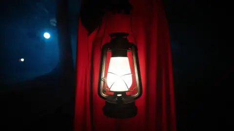 A red-cloaked figure carrying a lamp on The Traitors