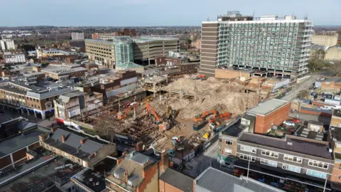 Capital&Centric An aerial drone shot shows the site surrounded by other buildings. The site has large mounds of dirt and gravel and there are orange construction vehicles within the site.