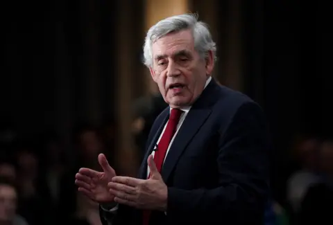 Gordon Brown wearing a white shirt and red tie address an unseen audience