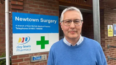 Andrew Turner/BBC Jonathan Knights is wearing a blue checked shirt and a blue round neck knitted jersey. He is standing outside the Newtown Surgery, where a sign states: "A branch of the East Norfolk Medical Practice. Telephone 01493 745050" and shows the logo and name of the Day Lewis pharmacy and the pharmacy green cross logo.