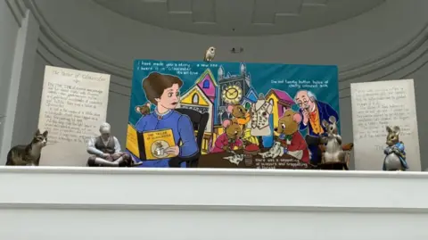 A display of figurines including a mice, a cat, a man and an owl. There is a large colourful painting behind it representing Beatrix Potter and her book The Tailor of Gloucester. There are two large plaques either side of the painting, with the words from the first and last page of the book on them. 