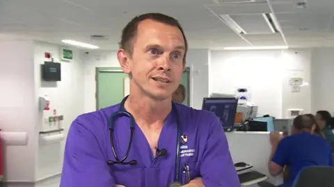 David Martin stands in the middle of the emergency department whil egiving an interview. He wears deep purple coloured scrubs, which have a logo on the chest saying 'NHS Emergency Department' and has a stethescope around his neck. He has short dark brown hair and his arms are crossed, he is smiling.