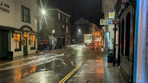 Kate Jones A view of flooding on a street. It is dark but is lit by street lamps. There is a lot of flood water on the road and there's pavements either side. 
