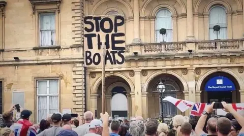 Hand holding up a banner reading 'Stop the boats' among a crowd of people, some waving Union and England flags, in front of a sand-coloured building