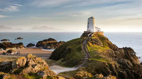 Getty Images View of Twr Mawr lighthouse on Angelsey surrounded by beach and large rocks on one side and the sea behind, and mountains in the background on the other side of the bay. the lighthouse is a circular white building with steps in the grassy rock on which it is perched leading up to it.