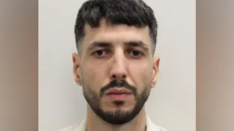Metropolitan Police Police custody mugshot of Brahim Keddad, a man with short dark hair, a trimmed dark beard and moustache, facing directly towards the camera against a plain off-white background.