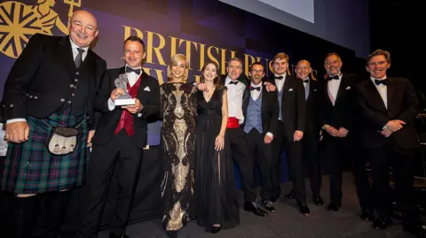 Dynisma A group of 10 people, two women and eight men, pose with their prize after winning an award at the British Business Awards. They are from the Bristol company Dynisma. The men are all in black tie and the women are in stylish dresses
