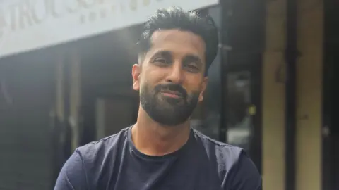 Aisha Iqbal/BBC A young man of South Asian heritage. He has a short, stylish beard. He wears a dark blue fitted t-shirt. He is standing in front of a shop.