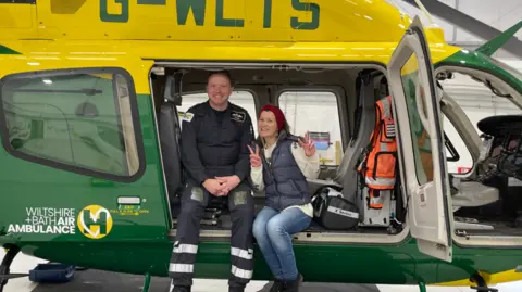 A woman doing peace signs while within a helicopter which says 'Wiltshire and Bath Air Ambulance'